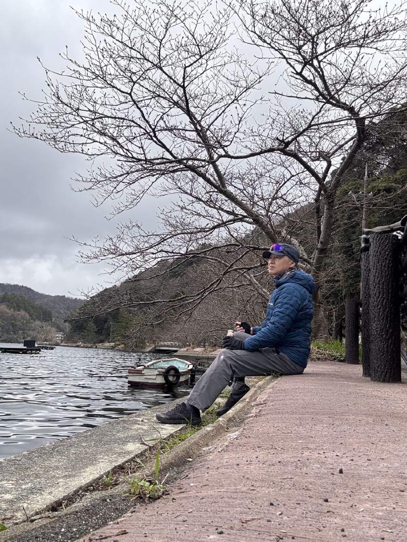 13:53,穿藍色外套的人坐在水邊的石岸上,後面是樹。
