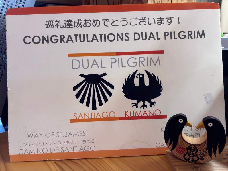15:28,熊野本宮館裡觀光協會的櫃台:「CONGRATULATIONS DUAL PILGRIM」,左邊是聖雅各的貝殼,右邊是熊野的八咫烏。