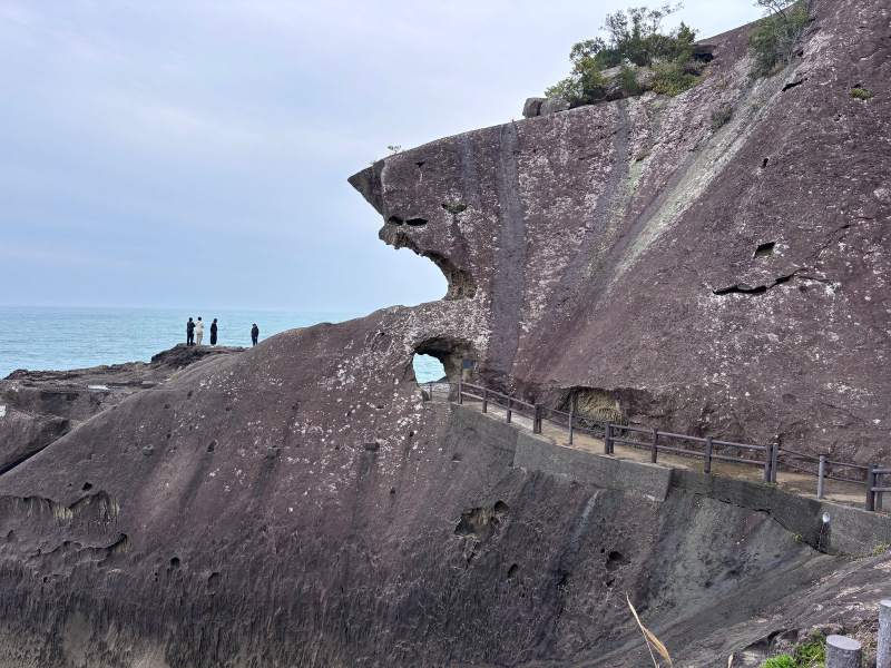 15:29,鬼城:整面岩壁滿是被海浪掏出的小洞,崖頭往海的方向伸出去,步道從底下的岩洞穿過;左邊站著的人小小的。